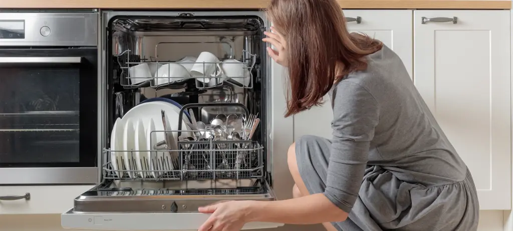 A woman is looking into an open dishwasher with clean, washed white dishes and stainless steel cutlery on a white, minimalist home kitchen. A convenient modern tool for household chores