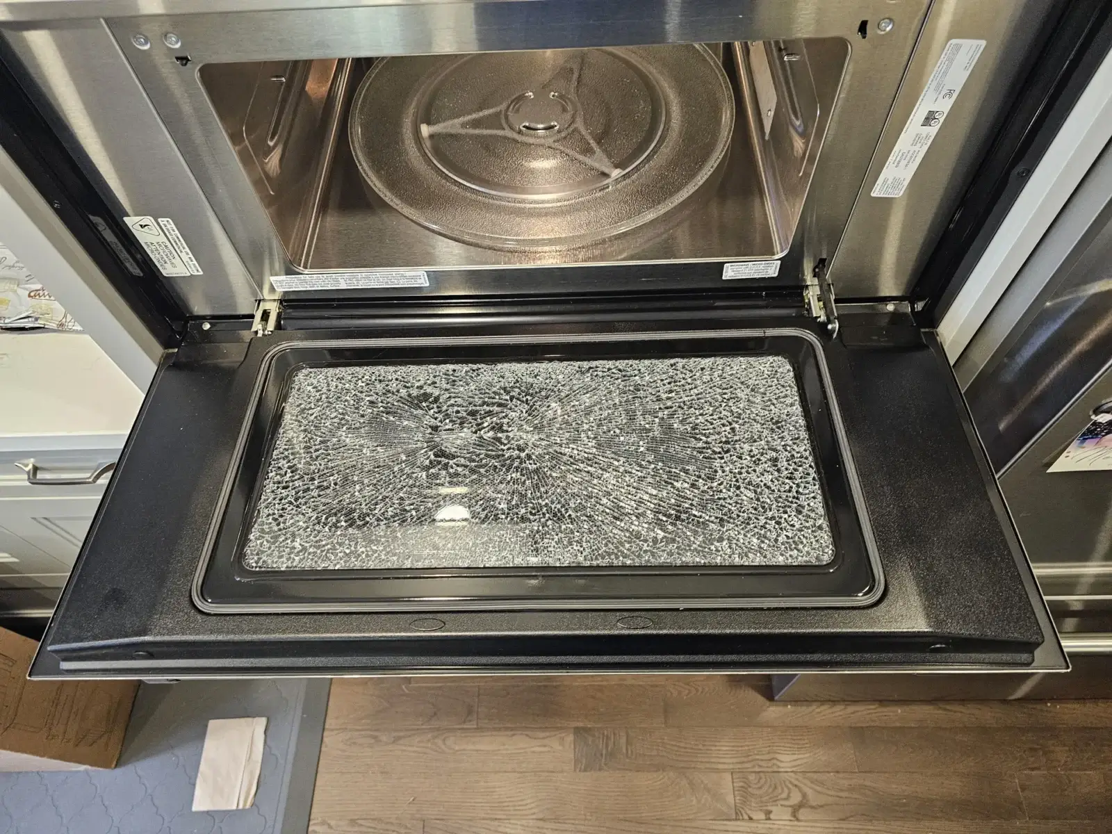 Damaged microwave with its drop-down door open and the door glass shattered but still held in the frame