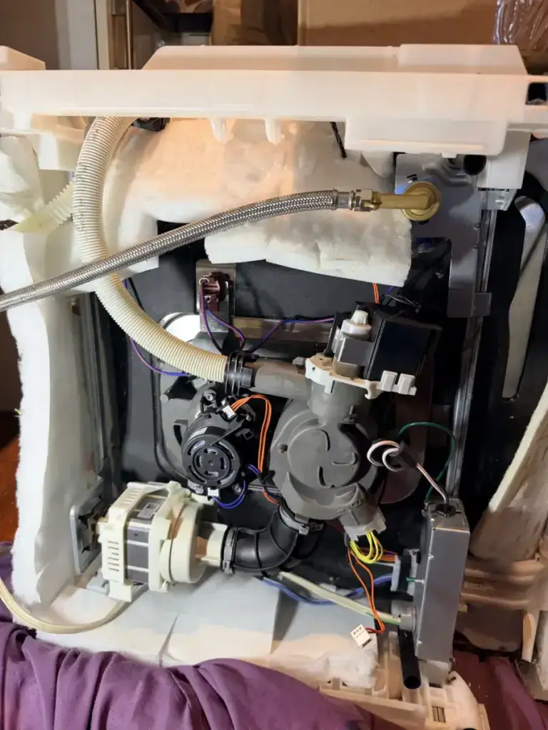 Internal view of a dishwasher with the pump, motor, and hoses exposed during a repair — the drain and circulation pumps are common failure points