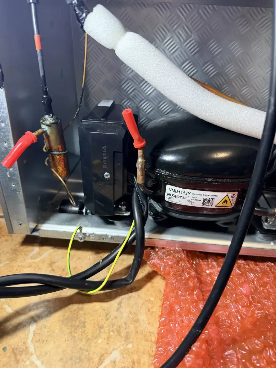 Black refrigerator compressor with its model label, a controller box beside it and two copper tube stubs capped with red plugs