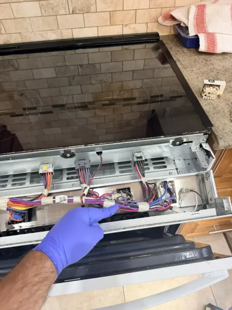 Gloved technician checking the colour-coded wiring harness beneath an open electric stove