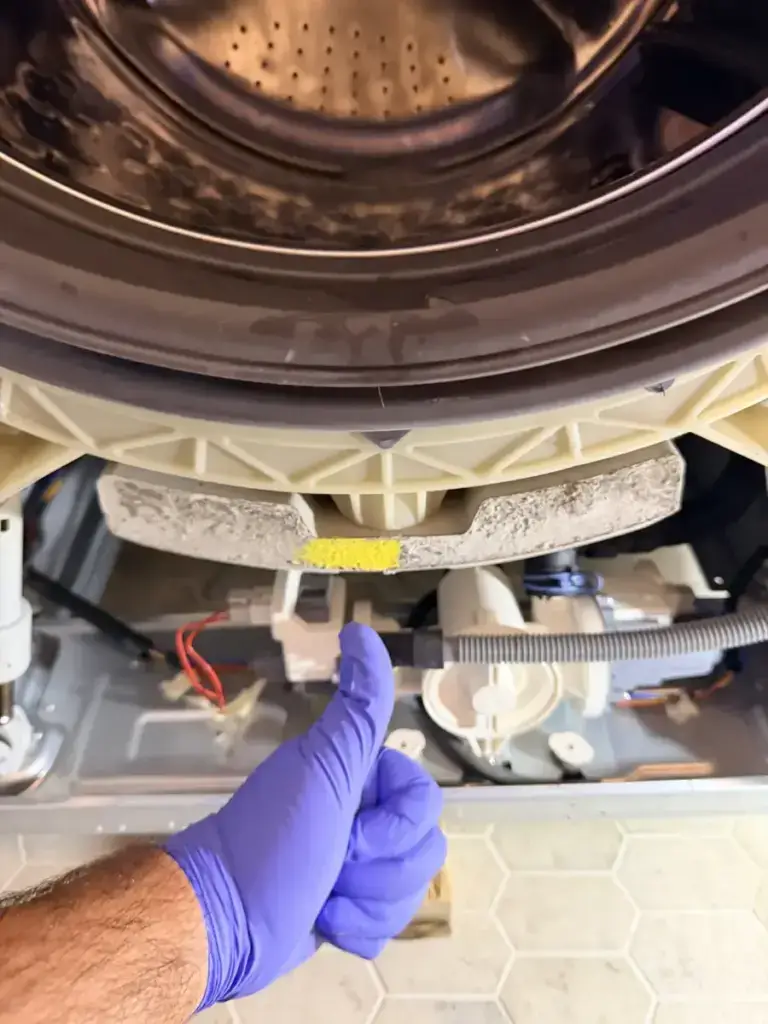 Gloved Appliance Forever technician giving a thumbs-up beside a disassembled front-load washer after a drain repair