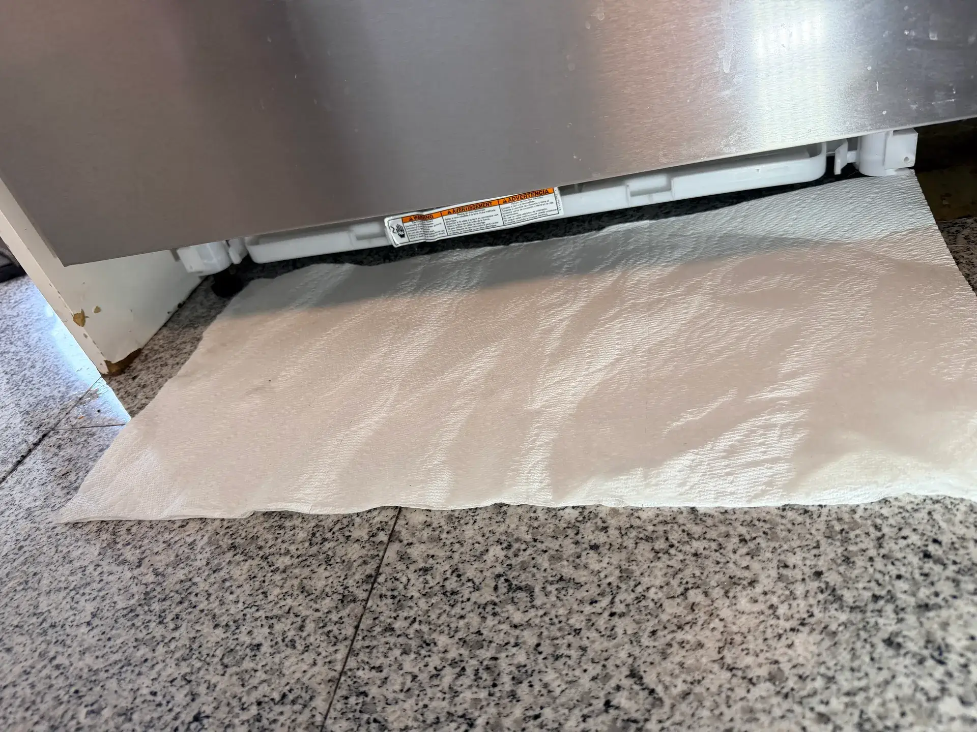 Paper towel laid under the front edge of a stainless steel dishwasher on the kitchen floor to show where a leak starts