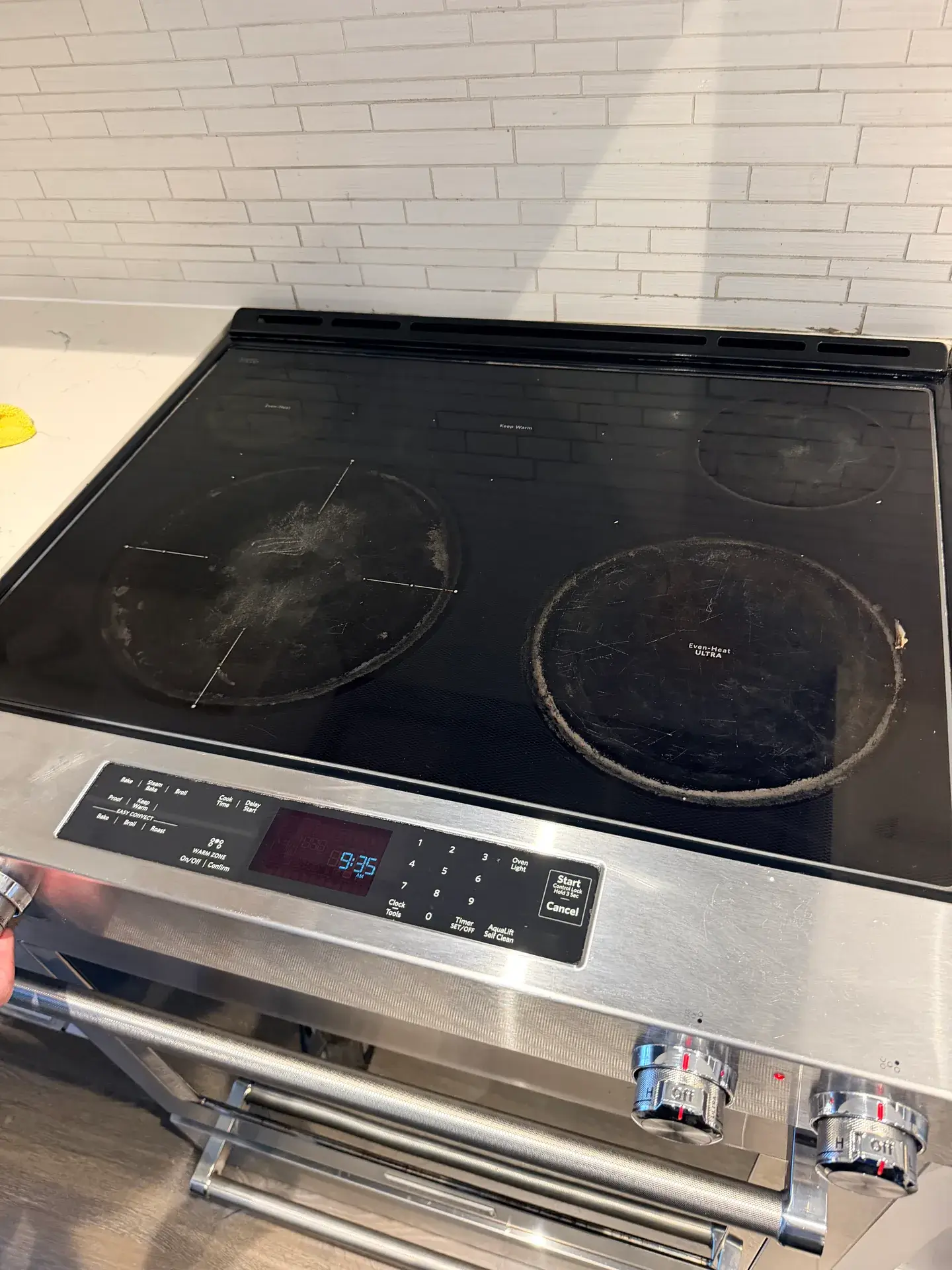 Black glass stove top with heavy grey rings and scratches on two cooking zones, control display lit, North York