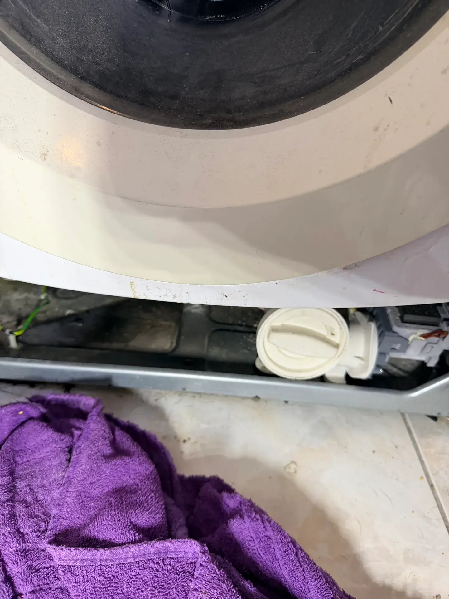 Round drain-pump filter cap at the bottom front of a front-load washer with a purple towel on the floor below