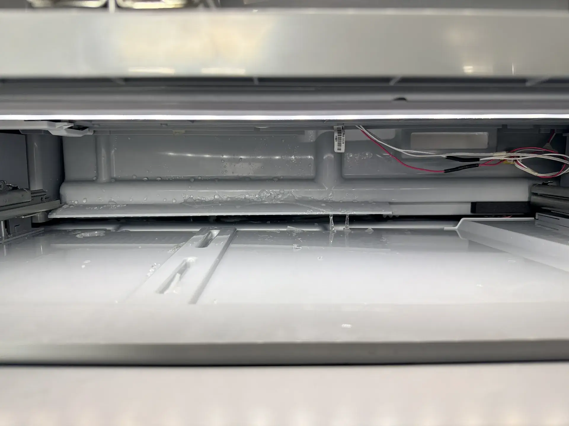 Refrigerator interior with the rear cover removed, showing a wet back wall and a red and white wiring harness