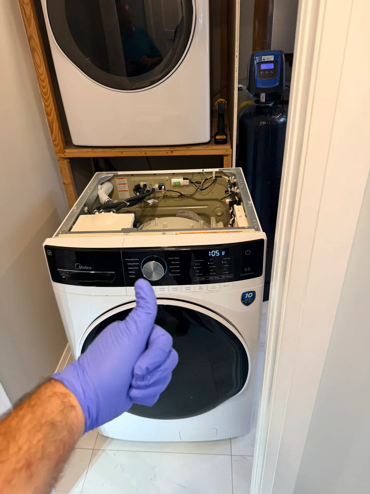 Midea front-load washer with its top panel removed exposing wiring and connectors, display lit, in an Innisfil laundry closet