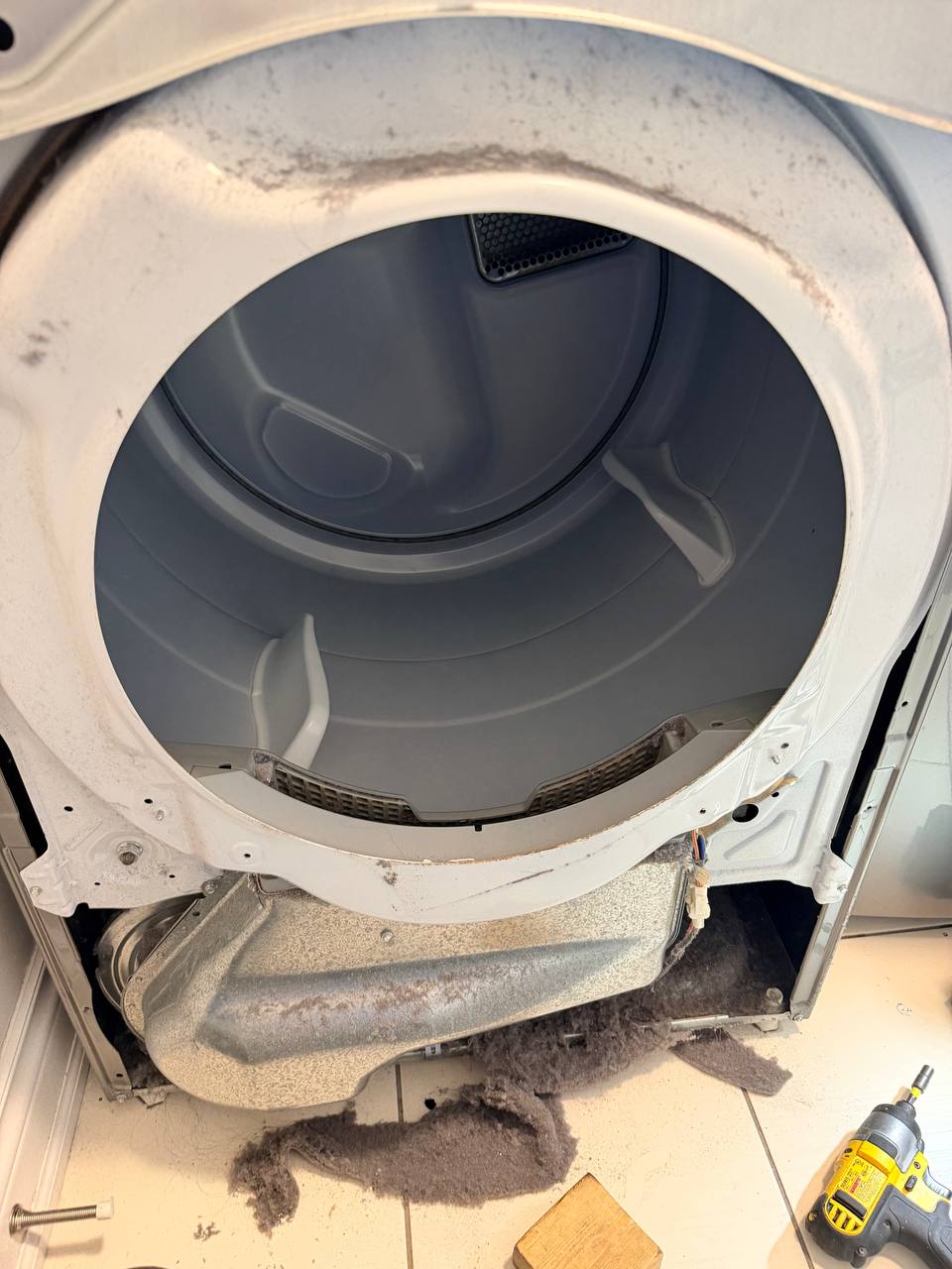 Dryer with the front panel off, grey lint on the front bulkhead and lower duct, and a pile of lint on the tile floor
