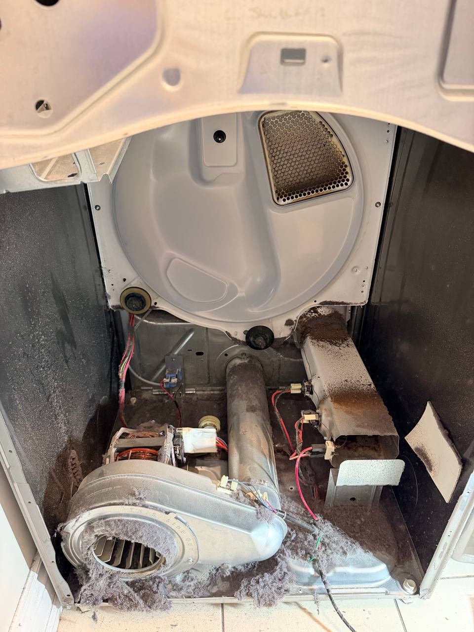 Wide view of the open dryer cabinet with lint over the floor pan, a packed blower inlet and a darkened heater duct